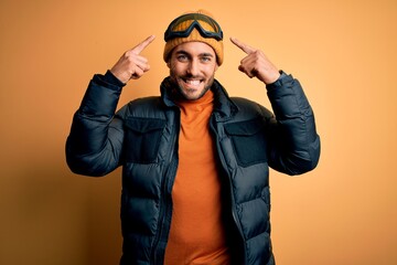 Young handsome skier man with beard wearing snow sportswear and ski goggles smiling pointing to head with both hands finger, great idea or thought, good memory
