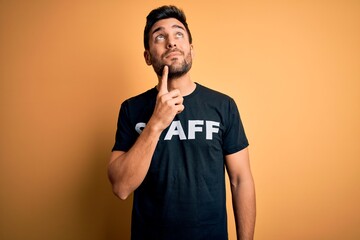 Young handsome worker man with beard wearing staff uniform t-shirt over yellow background serious face thinking about question, very confused idea