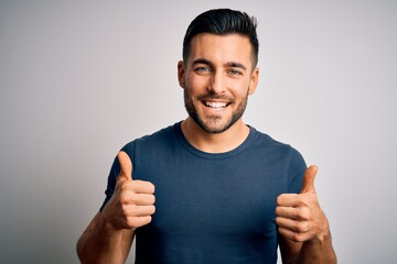 Young handsome man wearing casual t-shirt standing over isolated white background success sign doing positive gesture with hand, thumbs up smiling and happy. Cheerful expression and winner gesture.