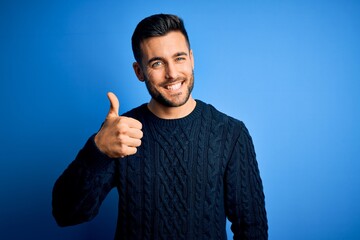 Young handsome man wearing casual sweater standing over isolated blue background doing happy thumbs...