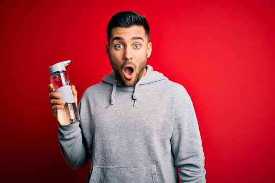 Young handsome sportsman drinking bottle of water to refeshment over red background scared in shock with a surprise face, afraid and excited with fear expression