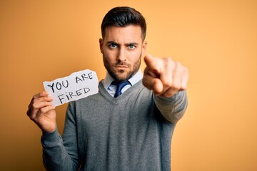 Young handsome man holding paper with you are fired message over yellow background pointing with...