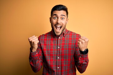 Young handsome man wearing casual shirt standing over isolated yellow background celebrating surprised and amazed for success with arms raised and open eyes. Winner concept.