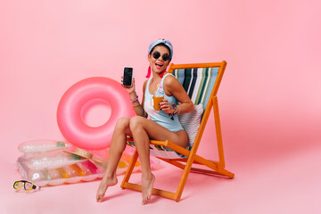 Cheerful girl in sunglasses sitting on deck chair with gadget. Gorgeous woman in swimsuit showing smartphone with blank screen.