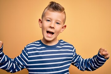 Young little caucasian kid with blue eyes wearing nautical striped shirt over yellow background celebrating surprised and amazed for success with arms raised and open eyes. Winner concept.