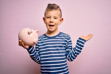 Young little caucasian kid banking money on piggy bank as wealth savings over pink background very happy and excited, winner expression celebrating victory screaming with big smile and raised hands