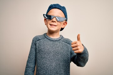 Young little caucasian kid wearing internet meme thug life glasses over isolated background doing happy thumbs up gesture with hand. Approving expression looking at the camera showing success.