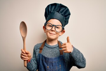 Young little caucasian cook kid wearing chef uniform and hat cooking using wooden spoon happy with...