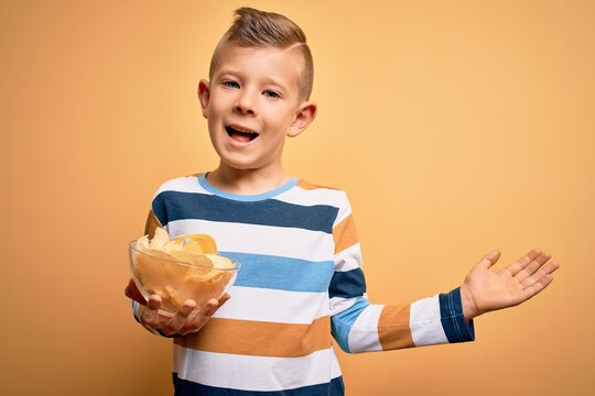 Young Little Caucasian Kid Eating Unheatlhy Potatoes Crisps Chips Over Yellow Background Very Happy And Excited, Winner Expression Celebrating Victory Screaming With Big Smile And Raised Hands