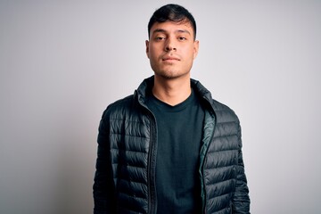 Young handsome hispanic man wearing winter coat standing over white isolated background with serious expression on face. Simple and natural looking at the camera.
