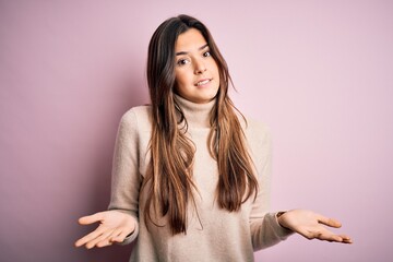 Young beautiful girl wearing casual turtleneck sweater standing over isolated pink background clueless and confused expression with arms and hands raised. Doubt concept.