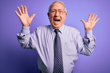 Grey haired senior business man wearing glasses standing over purple isolated background celebrating crazy and amazed for success with arms raised and open eyes screaming excited. Winner concept