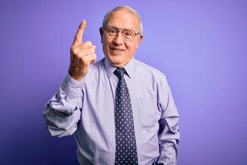 Grey haired senior business man wearing glasses standing over purple isolated background showing and pointing up with finger number one while smiling confident and happy.