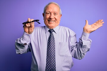 Senior grey haired man holding correction glasses over purple background very happy and excited,...