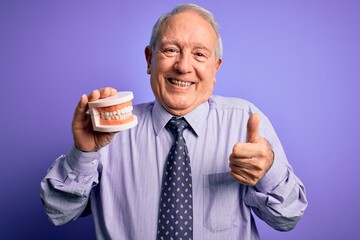 Grey haired senior man holding orthodontic prosthesis denture over purple background happy with big smile doing ok sign, thumb up with fingers, excellent sign