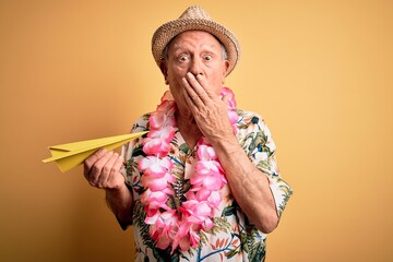 Grey haired senior man wearing summer hat and hawaiian lei holding paper plane on vacation cover mouth with hand shocked with shame for mistake, expression of fear, scared in silence, secret concept
