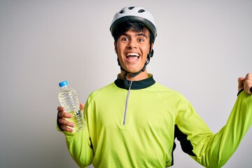 Young handsome cyclist man wearing security bike helmet drinking bottle of water screaming proud and celebrating victory and success very excited, cheering emotion