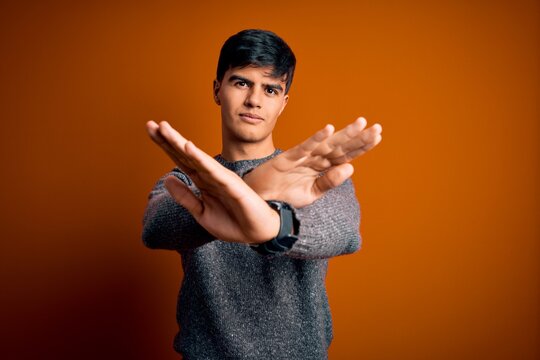Young handsome man wearing casual sweater standing over isolated orange background Rejection expression crossing arms and palms doing negative sign, angry face