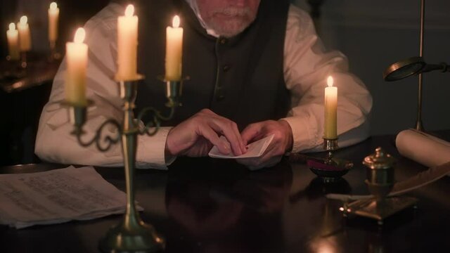 A scene from the 1800s era of a man sitting at a desk lit simply by candlelight who is opening a letter that has been sealed with a wax seal customary of the age.