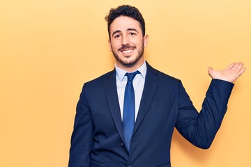 Young hispanic man wearing suit smiling cheerful presenting and pointing with palm of hand looking at the camera.
