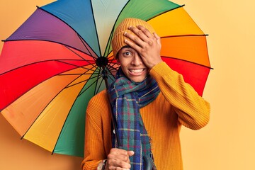 Young african amercian man holding colorful umbrella stressed and frustrated with hand on head,...
