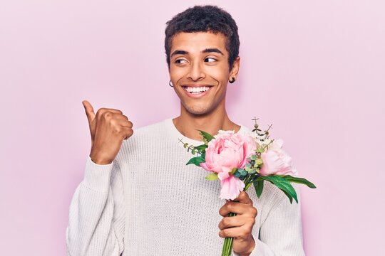 Young african amercian man holding flowers pointing thumb up to the side smiling happy with open mouth