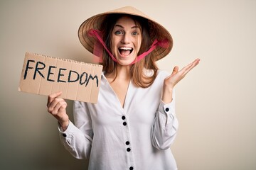 Young redhead woman wearing asian traditional hat holding banner with freedom message very happy and excited, winner expression celebrating victory screaming with big smile and raised hands