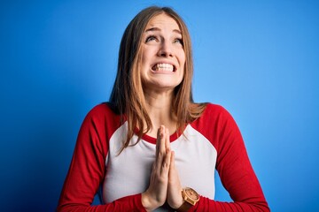 Young beautiful redhead woman wearing casual t-shirt over isolated blue background begging and...