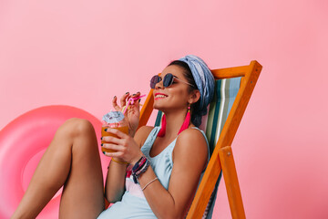 Dreamy woman in sunglasses drinking cocktail. Studio shot of girl in swimsuit lying in deck chair on pink background. - Powered by Adobe