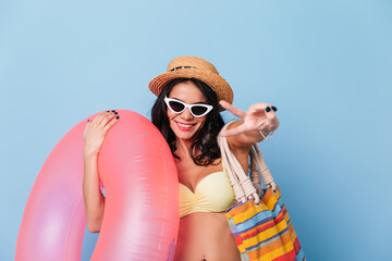 Cute tanned girl in sunglasses showing peace sign. Studio shot of positive lady holding swimming circle with smile.