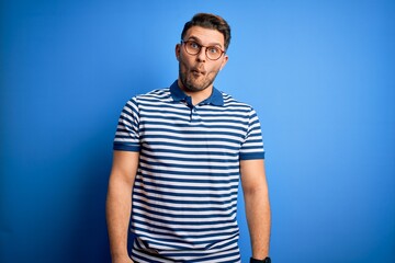 Obraz premium Young man with blue eyes wearing glasses and casual striped t-shirt over blue background making fish face with lips, crazy and comical gesture. Funny expression.
