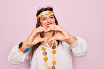 Young hispanic hippie woman wearing fashion boho style and sunglasses over pink background smiling in love doing heart symbol shape with hands. Romantic concept.