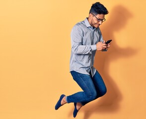 Fototapeta na wymiar Young handsome latin man wearing glasses jumping with smile on face. Using smartphone over isolated yellow background