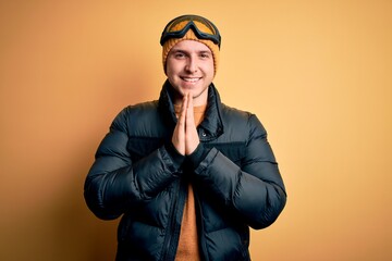 Young handsome caucasian man wearing hat, coat and ski glasses for winter and snow weather praying with hands together asking for forgiveness smiling confident.