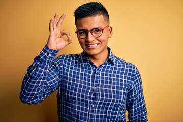 Young handsome latin man wearing casual shirt and glasses over yellow background smiling positive doing ok sign with hand and fingers. Successful expression.