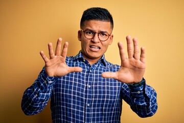 Young handsome latin man wearing casual shirt and glasses over yellow background afraid and terrified with fear expression stop gesture with hands, shouting in shock. Panic concept.