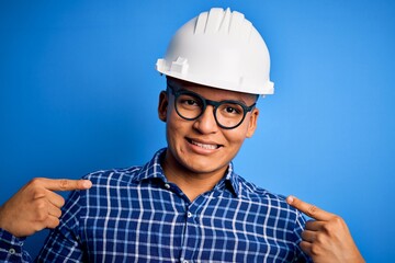 Young handsome engineer latin man wearing safety helmet over isolated blue background looking confident with smile on face, pointing oneself with fingers proud and happy.