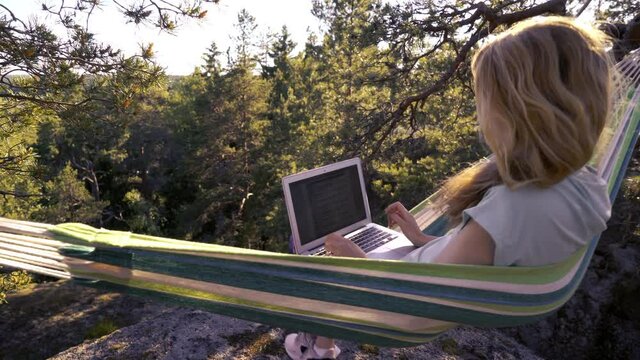 A young woman working on a laptop while lying in a hammock in the woods. Self-isolation, freelancing, remote work