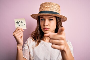 Young blonde girl wearing a hat holding paper note with yes word as positive message pointing with...