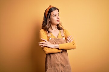 Obraz premium Young beautiful blonde girl wearing overall standing over yellow isolated background looking to the side with arms crossed convinced and confident