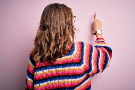 Young beautiful blonde girl wearing glasses and casual sweater over pink isolated background Posing backwards pointing ahead with finger hand