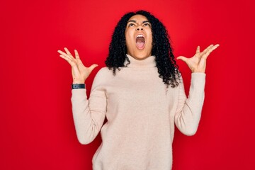 Young african american curly woman wearing casual turtleneck sweater over red background crazy and mad shouting and yelling with aggressive expression and arms raised. Frustration concept.
