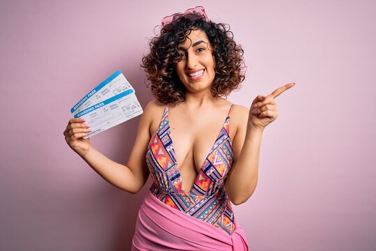 Young beautiful tourist arab woman on vacation wearing swimsuit holding boarding pass airline very happy pointing with hand and finger to the side