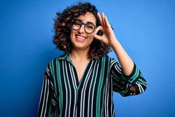 Young beautiful curly arab woman wearing striped shirt and glasses over blue background smiling positive doing ok sign with hand and fingers. Successful expression.