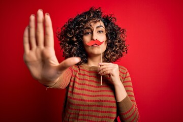 Young beautiful curly arab woman holding funny mustache over isolated red background with open hand...