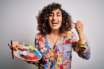 Young beautiful curly arab artist woman painting using brush and palette with colors screaming proud and celebrating victory and success very excited, cheering emotion - obrazy, fototapety, plakaty