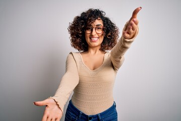 Young beautiful curly arab woman wearing casual t-shirt and glasses over white background looking at the camera smiling with open arms for hug. Cheerful expression embracing happiness.