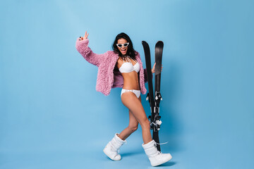 Blithesome brunette girl in bikini and jacket dancing on blue background. Full length view of pretty young woman with skis.