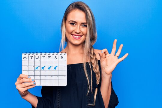 Young beautiful blonde tourist woman holding travel calendar showing vacation week doing ok sign with fingers, smiling friendly gesturing excellent symbol