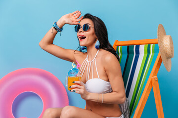 Emotional girl in bikini holding glass of juice and looking away. Studio shot of blissful brunette lady in sunglasses sitting in chaise longue.